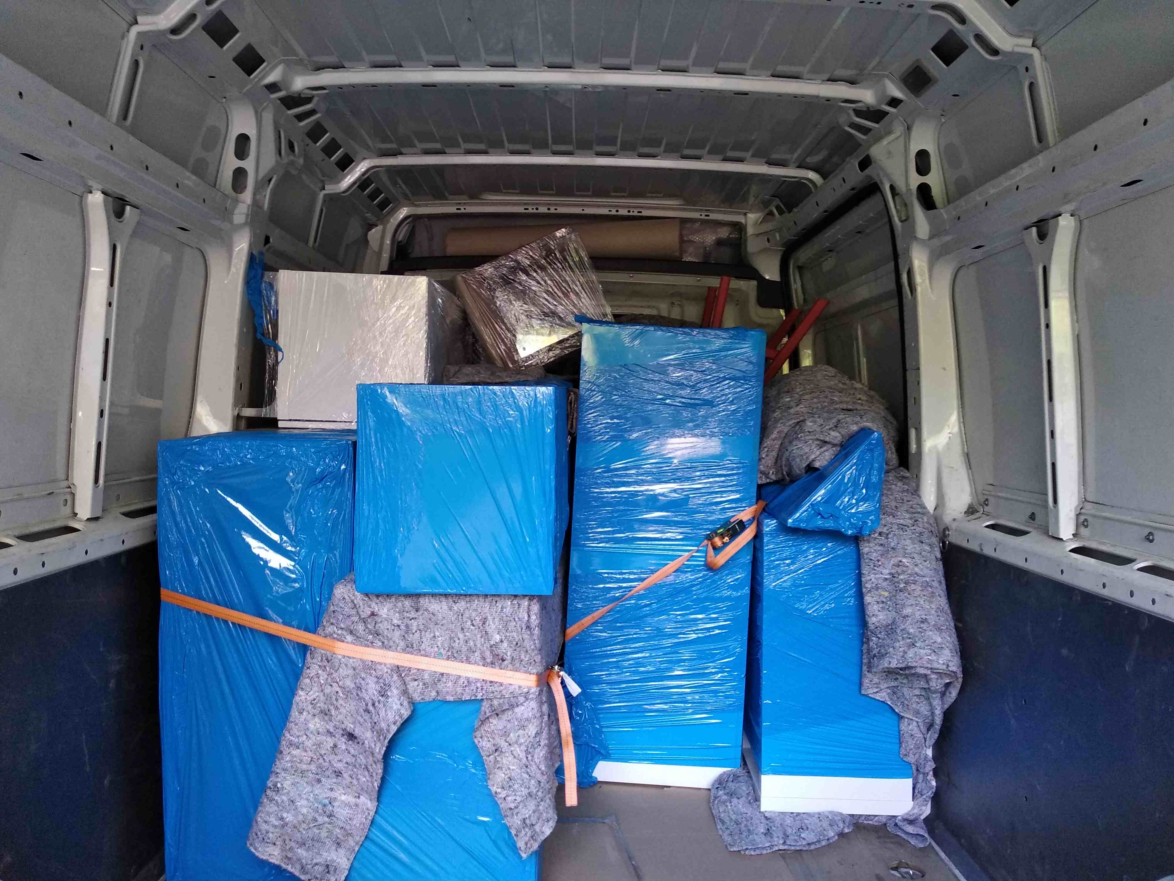 Wnętrze furgonetki wypełnionej zabezpieczonymi paczkami i meblami do przeprowadzki we Wrocławiu, owiniętymi w niebieską folię i szare koce, zabezpieczone pomarańczowymi pasami.