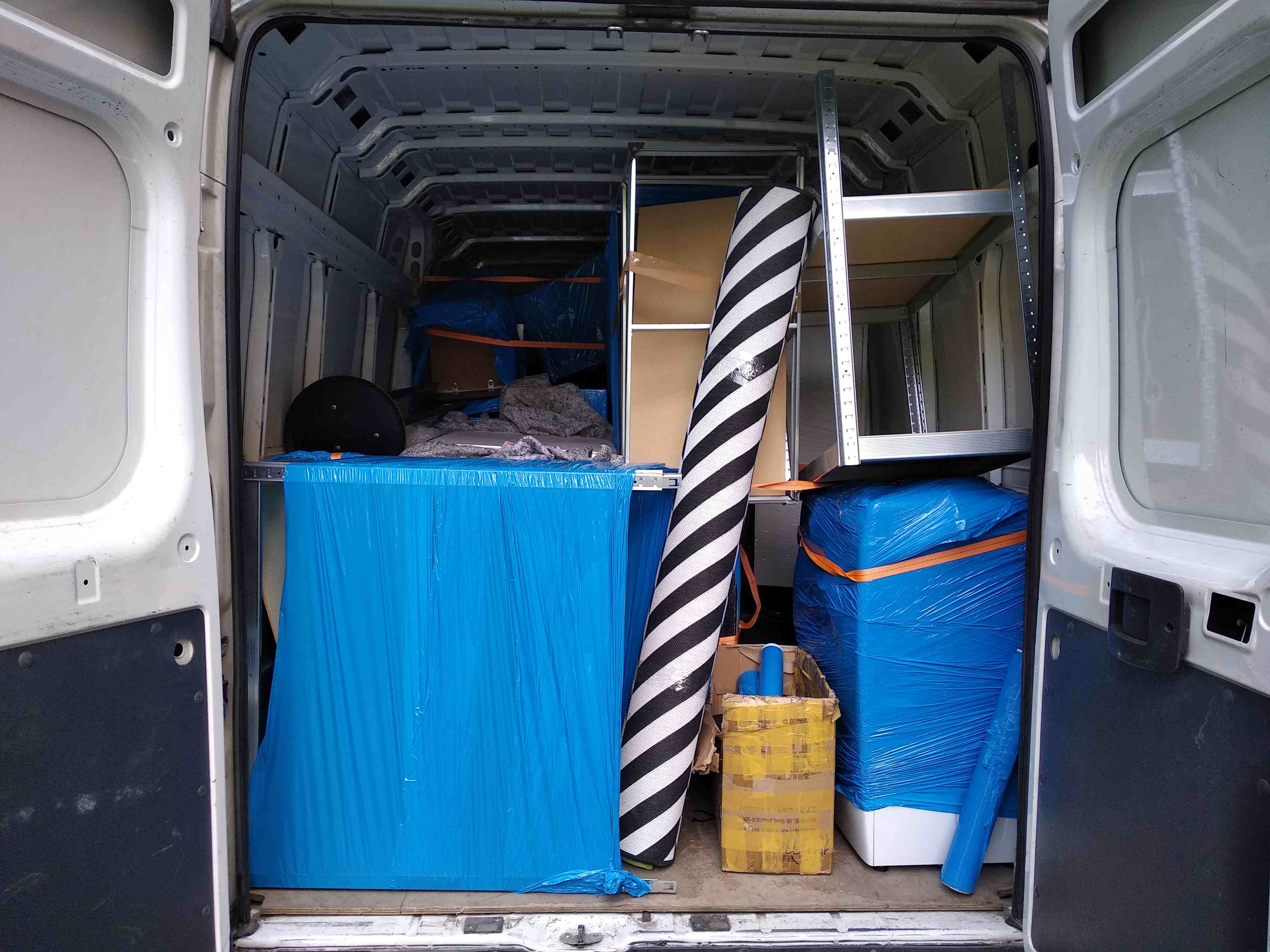 Wnętrze furgonetki załadowane meblami i przedmiotami zabezpieczonymi folią, gotowymi do transportu. Widoczny zwinięty dywan w biało-czarne pasy oraz metalowy regał.