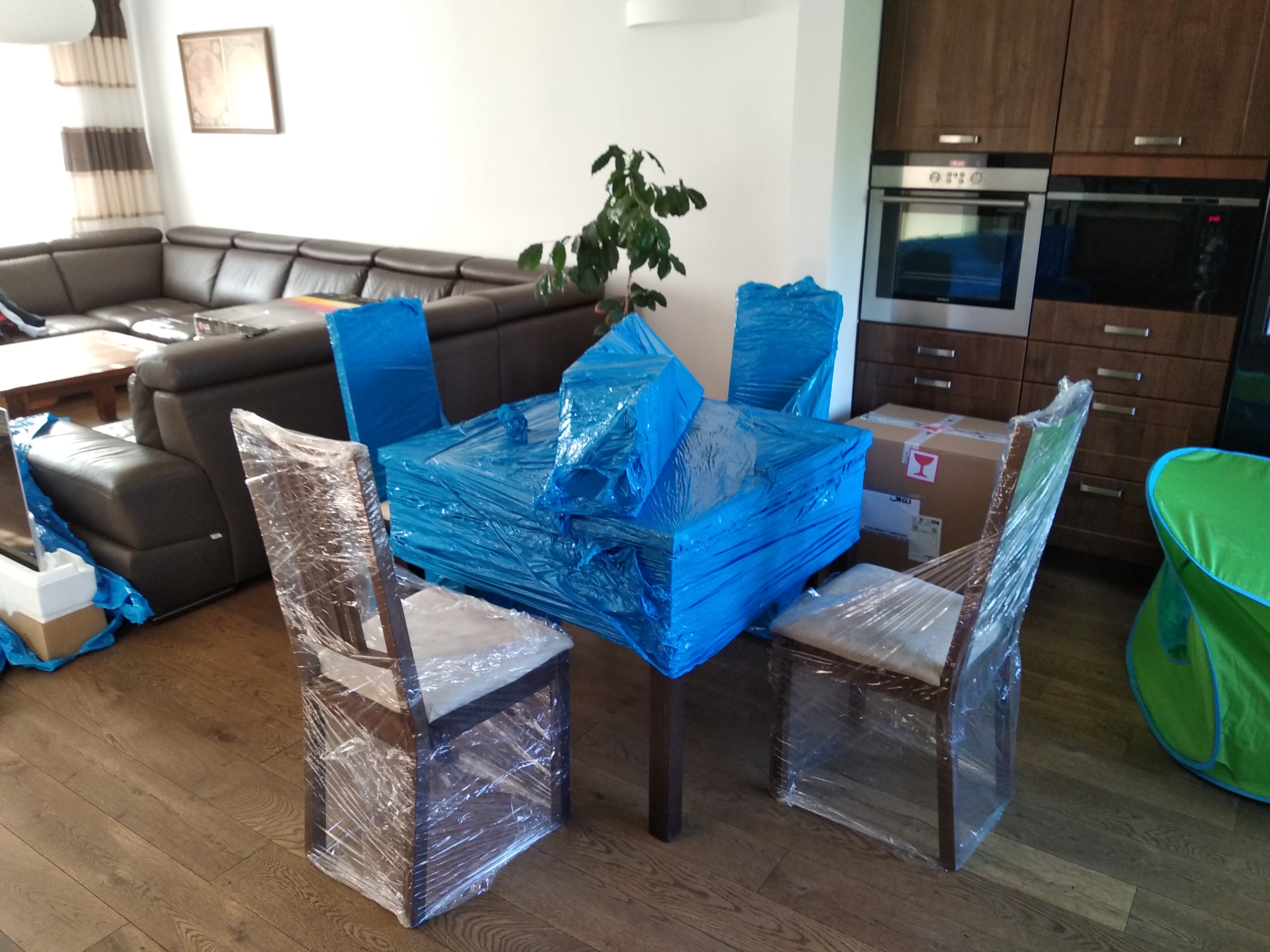 Stół i krzesła owinięte folią stretch oraz niebieską folią ochronną w trakcie przygotowań do transportu, widoczny narożnik skórzanej sofy w tle