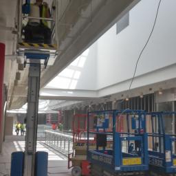 SORPLEX Sp. z o.o. - Pracownik w żółtej kamizelce i kasku na podnośniku koszowym, montujący instalacje pod sufitem w nowym budynku z dużymi oknami dachowymi. Widoczne koryta kablowe i inne podnośniki. W tle inni...