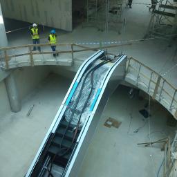 SORPLEX Sp. z o.o. - Widok z góry na montaż ruchomych schodów w budynku w stanie surowym; pracownicy w kaskach i kamizelkach odblaskowych obserwują zawieszone schody ruchome, zabezpieczone folią.