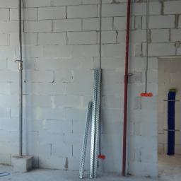 SORPLEX Sp. z o.o. - Surowy stan instalacji elektrycznej w nowo wzniesionym budynku z bloków betonu komórkowego, widoczne przewody elektryczne zwisające ze ściany, rurki ochronne oraz pionowa rura instalacyjna z uchwytem.