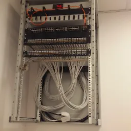 Szafa rack z okablowaniem sieciowym w biurze, widoczne patch panele, switche i uporządkowane kable kategorii 5e/6, zasilacz awaryjny w dolnej części szafy.