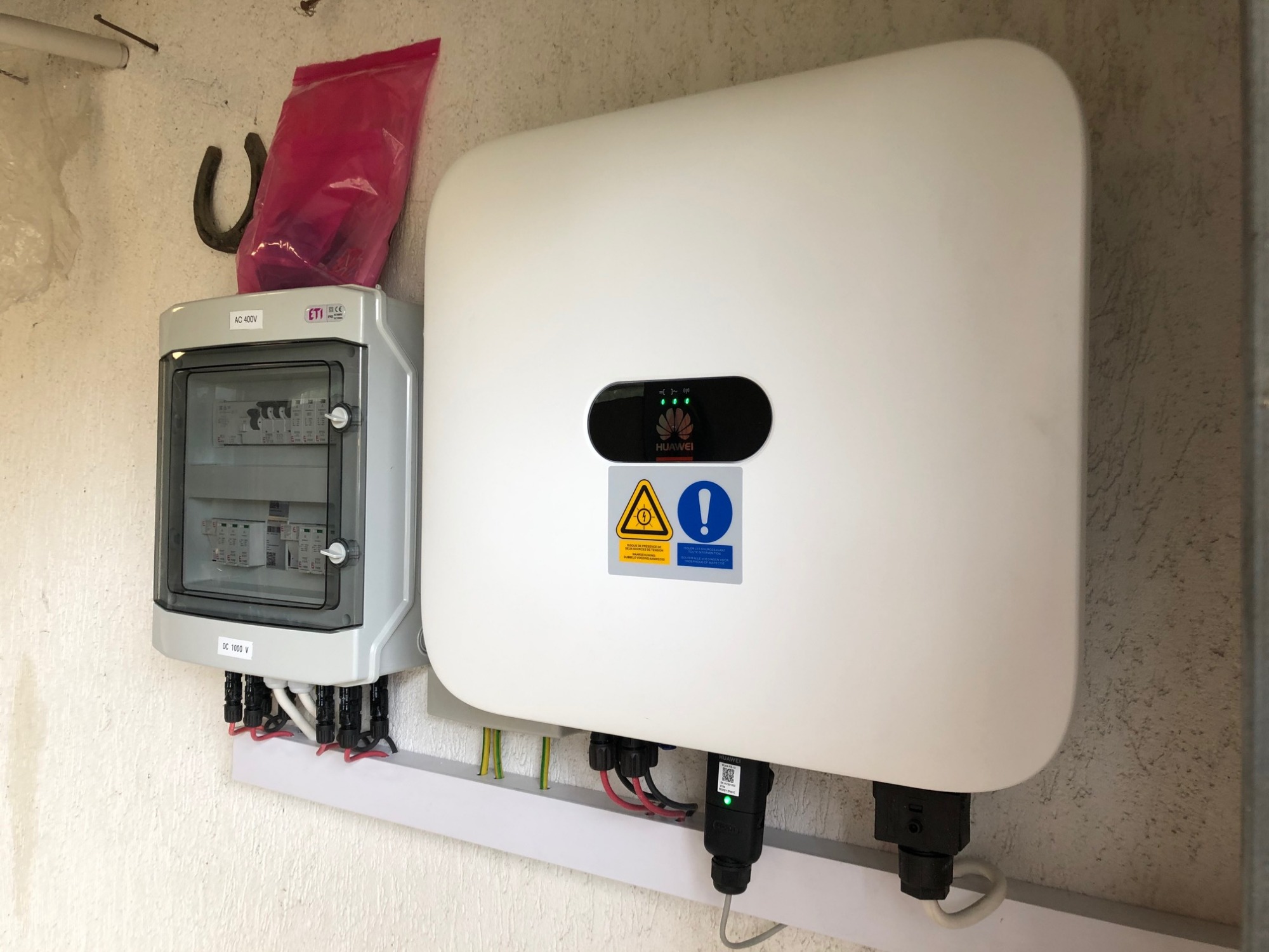 Biały falownik marki Huawei zamontowany na ścianie obok skrzynki elektrycznej z bezpiecznikami, widoczne przewody i elementy instalacji.