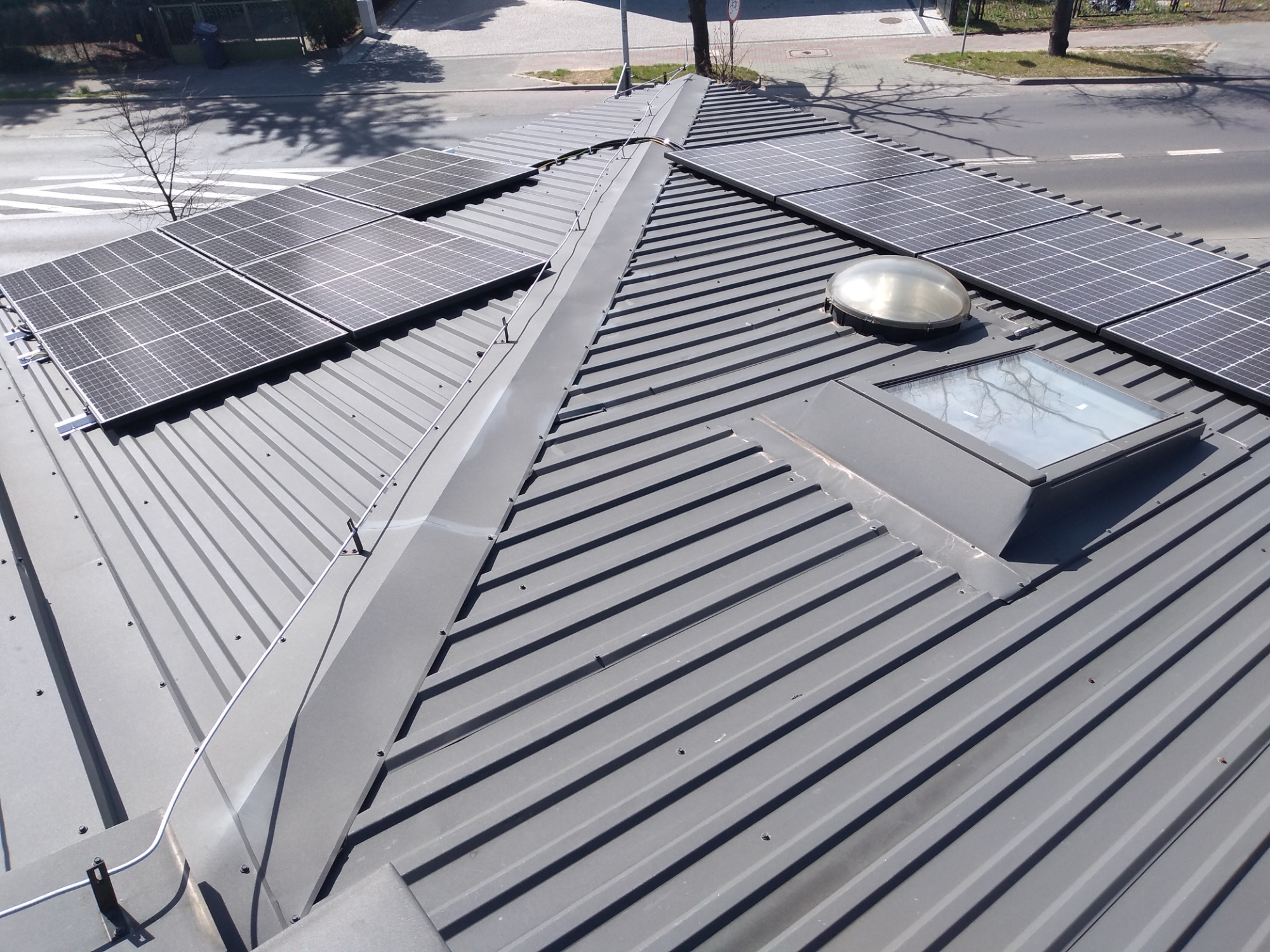 Panele fotowoltaiczne zamontowane na szarym, metalowym dachu z blachy trapezowej, widoczne okno dachowe i świetlik kopułkowy, ulica w tle.