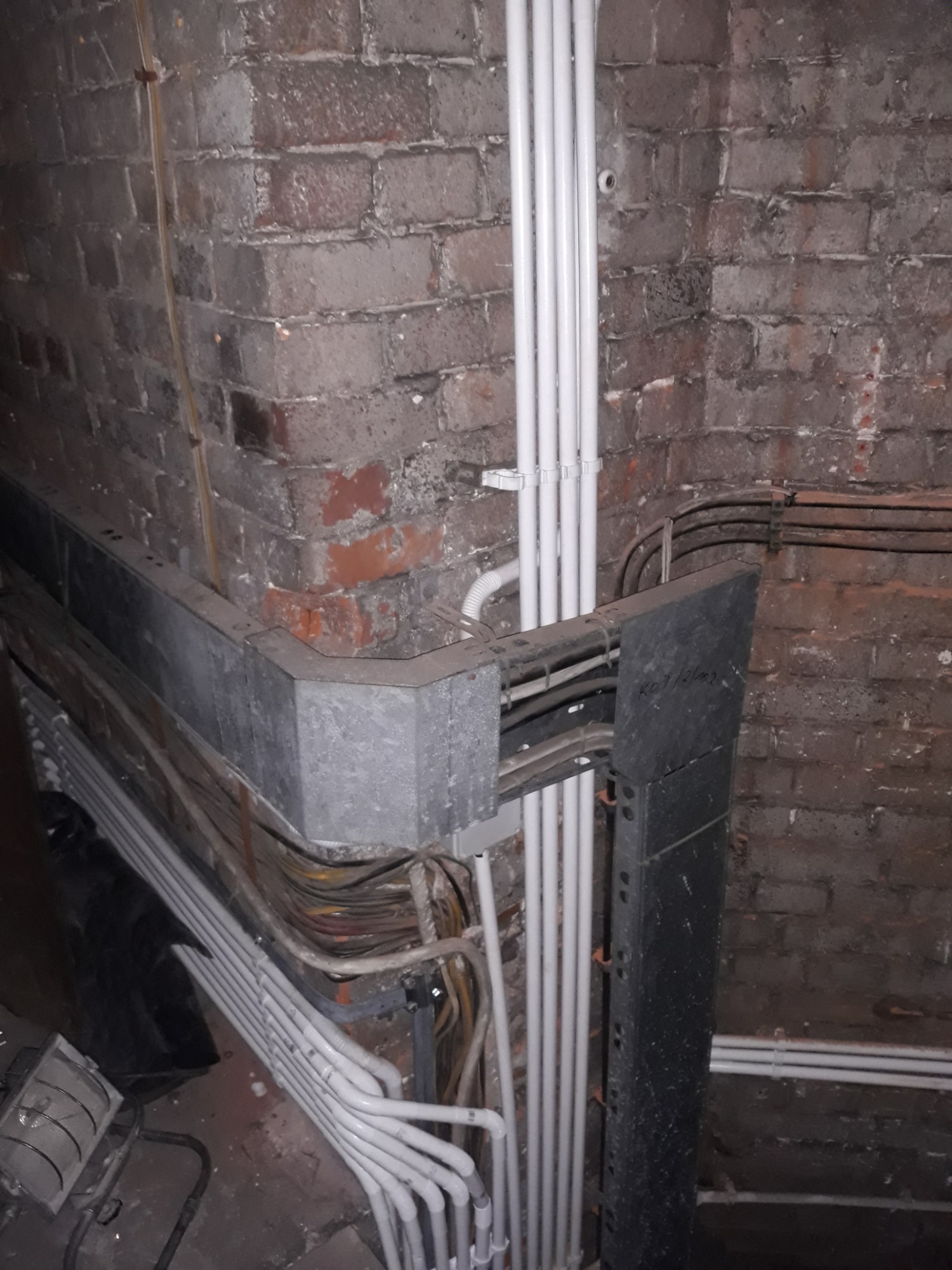 Kąt piwnicy z odsłoniętą cegłą, gdzie widoczne są rury PCV prowadzone pionowo wzdłuż ściany, metalowa korytka kablowa z przewodami oraz metalowa konstrukcja wsporcza.