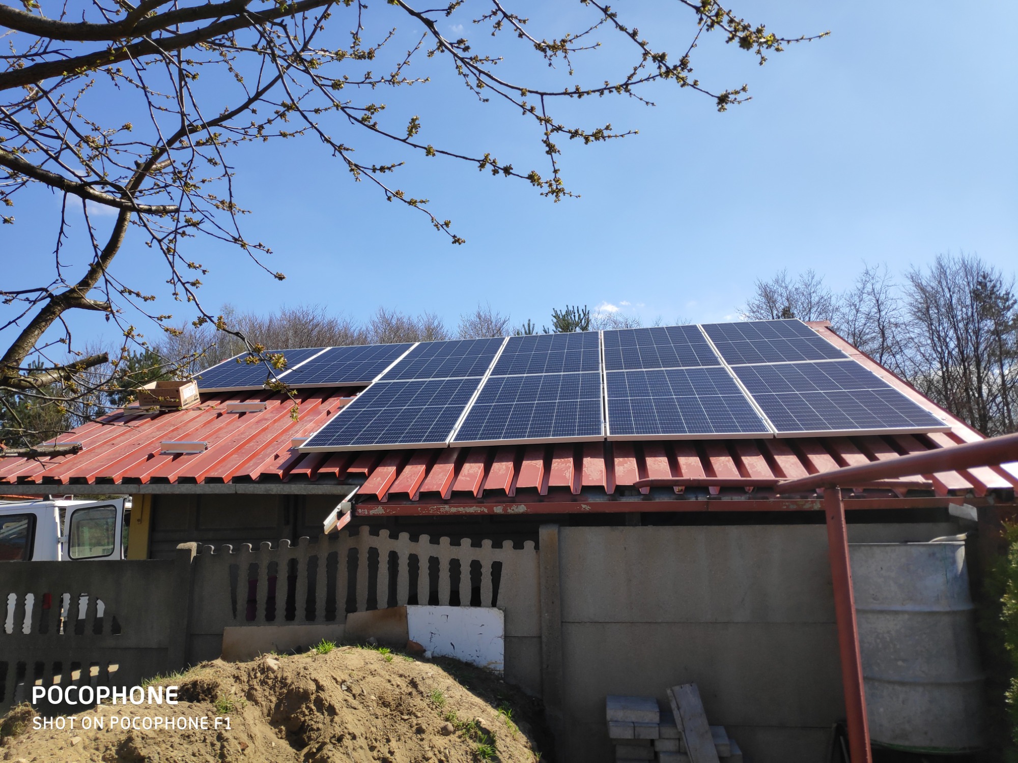 Panele słoneczne na czerwonym, blaszanym dachu domu jednorodzinnego, widoczne z poziomu gruntu, z gałęziami drzewa w kwitnącymi pąkami na pierwszym planie i fragmentem białego samochodu dostawczego...