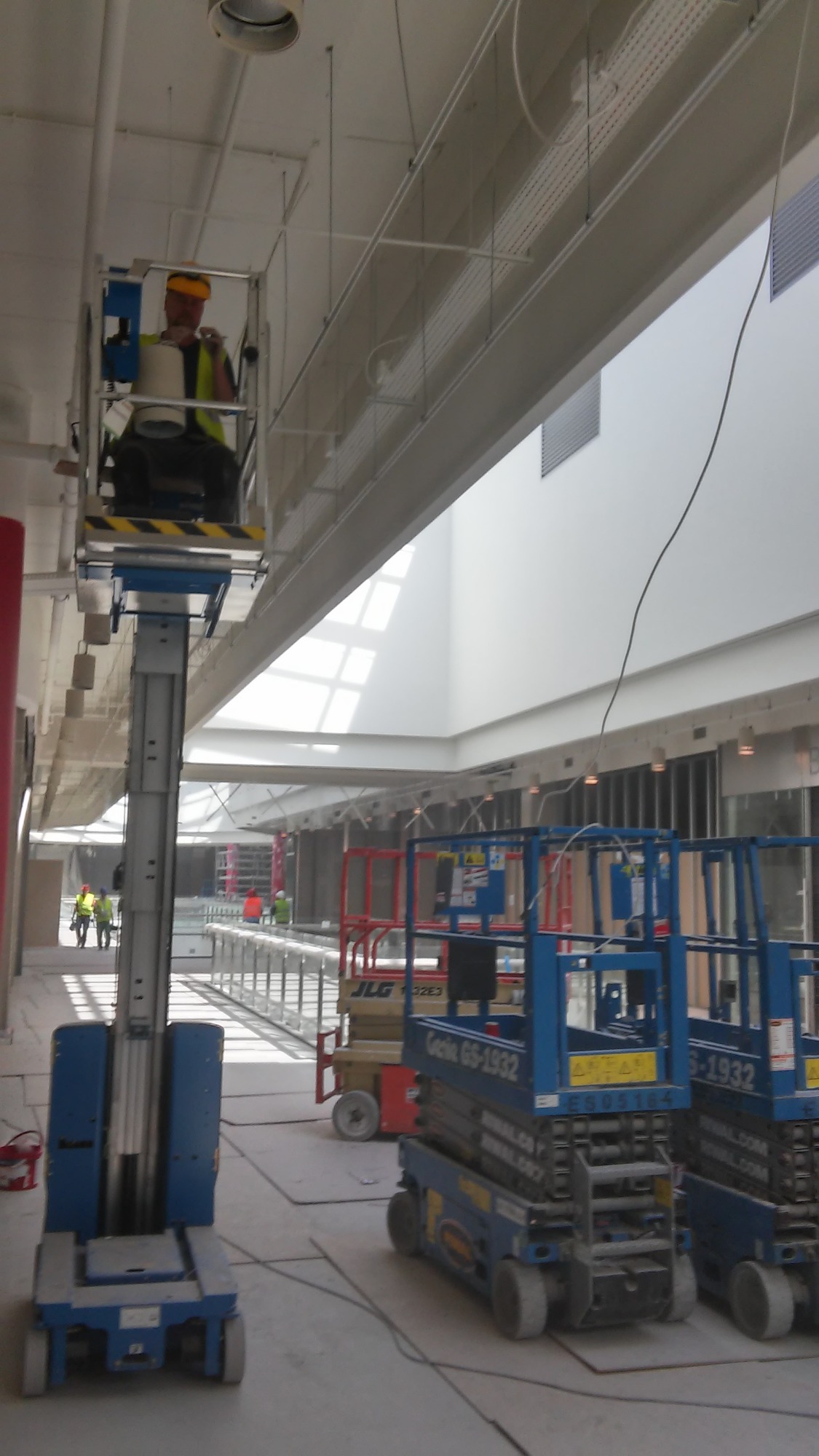 Pracownik w żółtej kamizelce i kasku na podnośniku koszowym, montujący instalacje pod sufitem w nowym budynku z dużymi oknami dachowymi. Widoczne koryta kablowe i inne podnośniki. W tle inni...