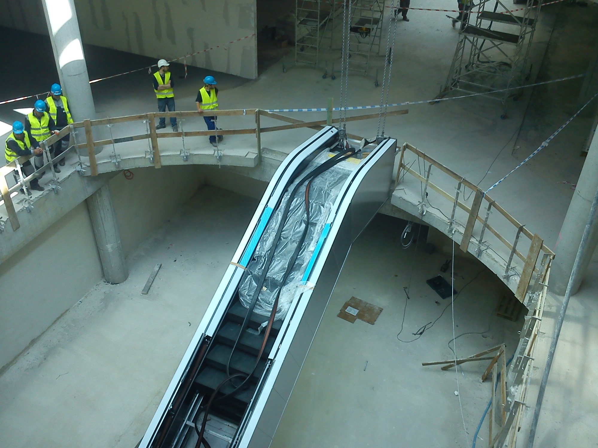 Widok z góry na montaż ruchomych schodów w budynku w stanie surowym; pracownicy w kaskach i kamizelkach odblaskowych obserwują zawieszone schody ruchome, zabezpieczone folią.
