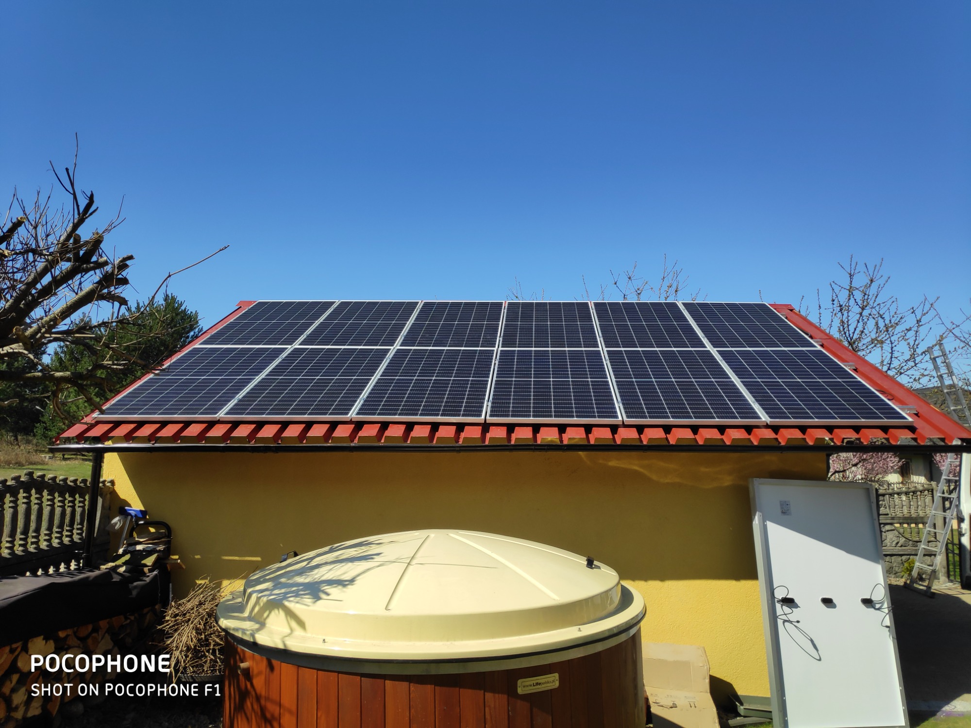 Panele słoneczne na dachu budynku z czerwoną dachówką, widoczne również jacuzzi ogrodowe z drewnianą obudową i niezamontowany panel słoneczny oparty o ścianę.