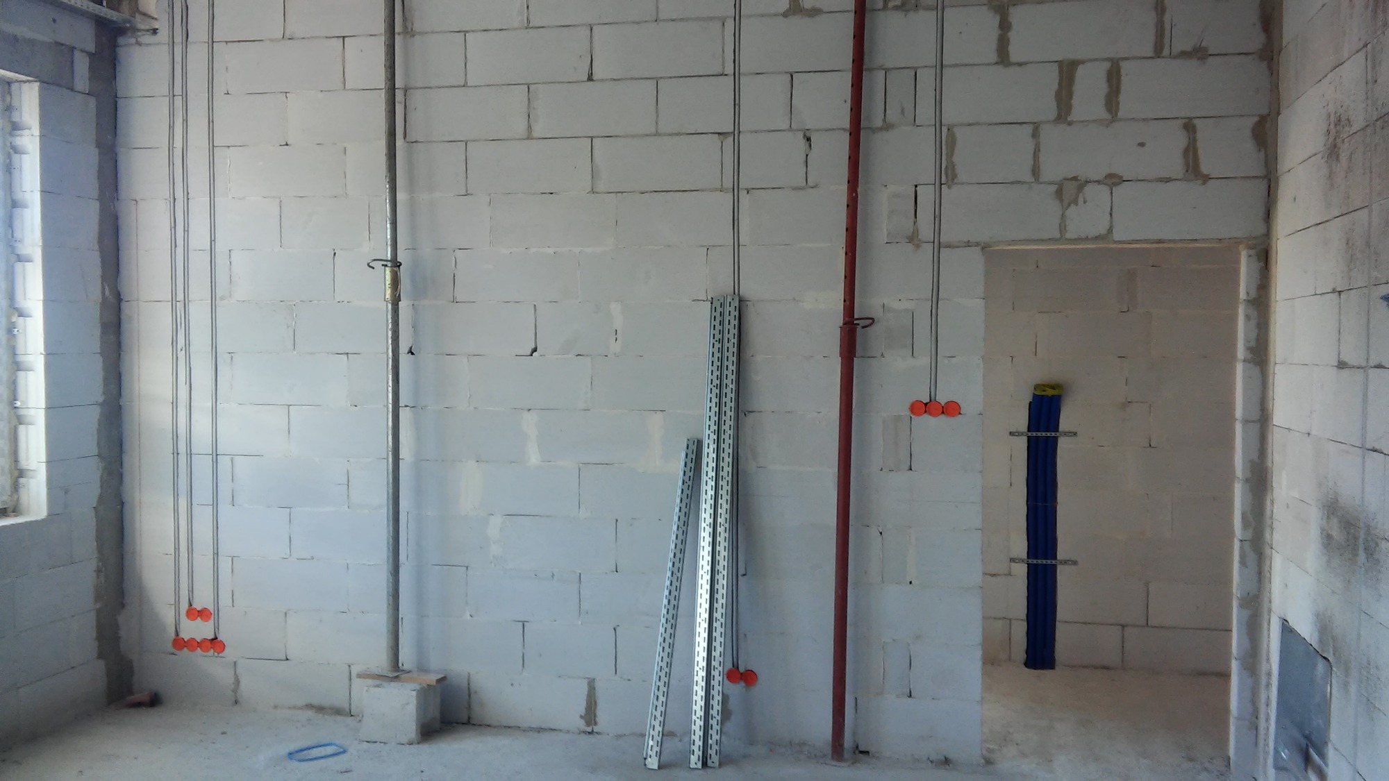 Surowy stan instalacji elektrycznej w nowo wzniesionym budynku z bloków betonu komórkowego, widoczne przewody elektryczne zwisające ze ściany, rurki ochronne oraz pionowa rura instalacyjna z uchwytem.
