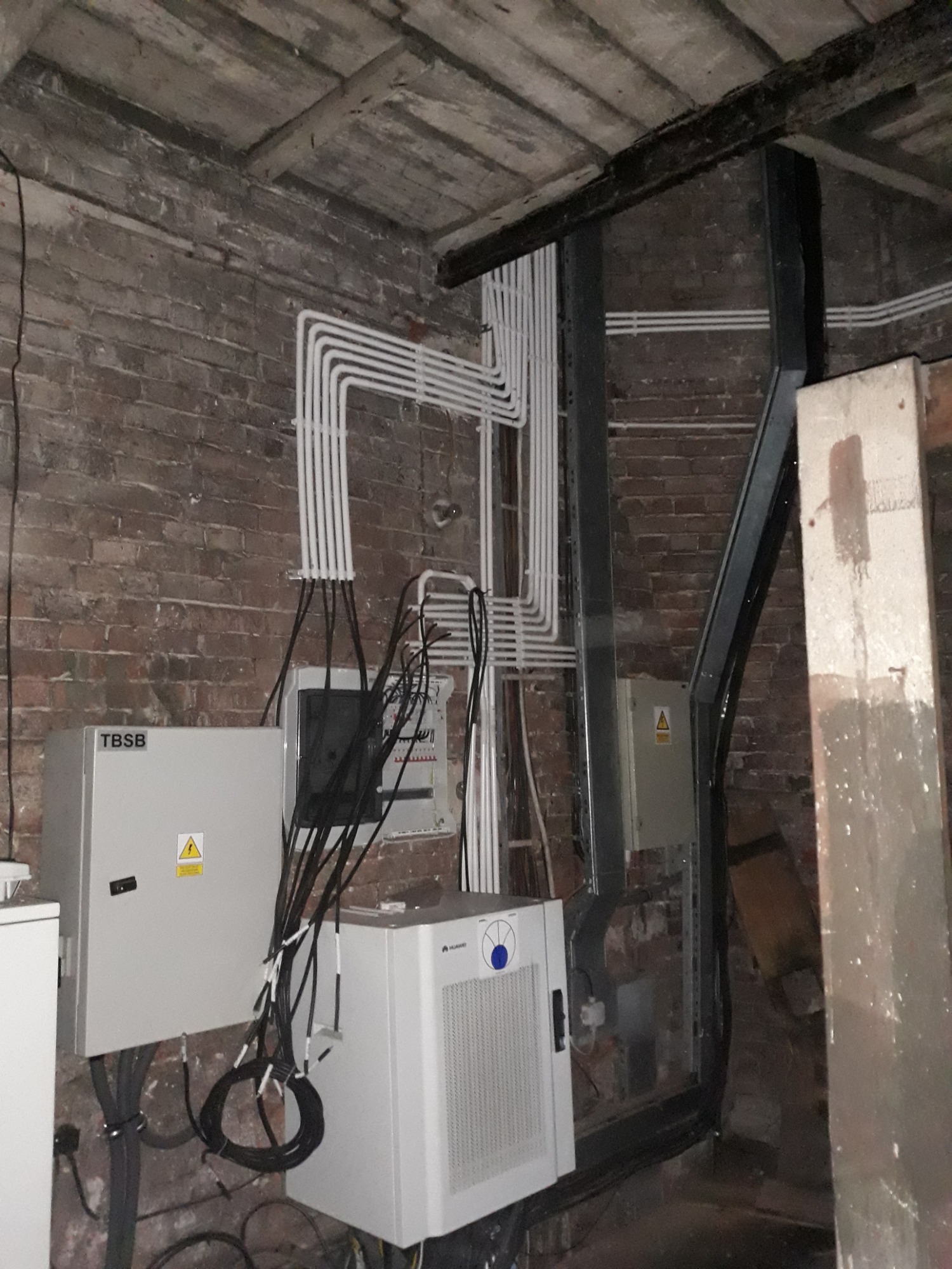 Szare skrzynki elektryczne TBSB z żółtymi znakami ostrzegawczymi, liczne białe kable w peszlach na tle starej ceglanej ściany i drewnianego stropu w piwnicy.