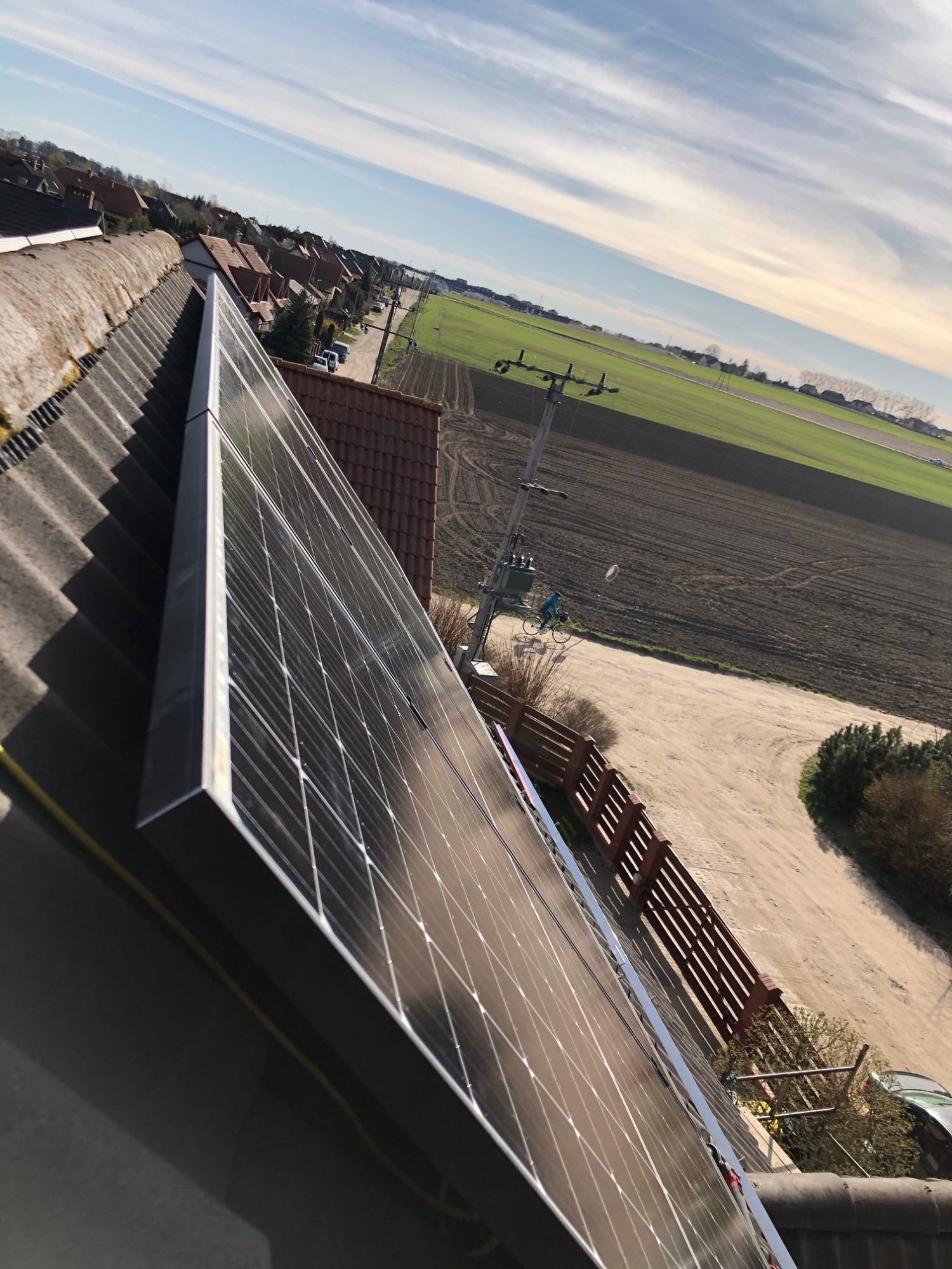 Panel słoneczny zamontowany na dachu z widokiem na wiejską okolicę, rowerzysta na polnej drodze w tle.