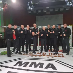 Grupa mężczyzn w czarnych strojach, prawdopodobnie pracownicy ochrony, pozuje na ringu MMA z logo MD Domy z Drewna, w tle siatka ringu i oświetlenie sceniczne.