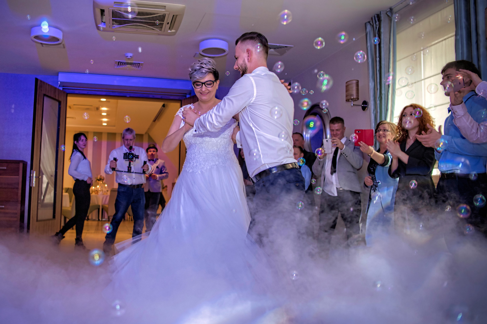 Para młoda tańczy w dymie i bańkach mydlanych, otoczona gośćmi weselnymi robiącymi zdjęcia.