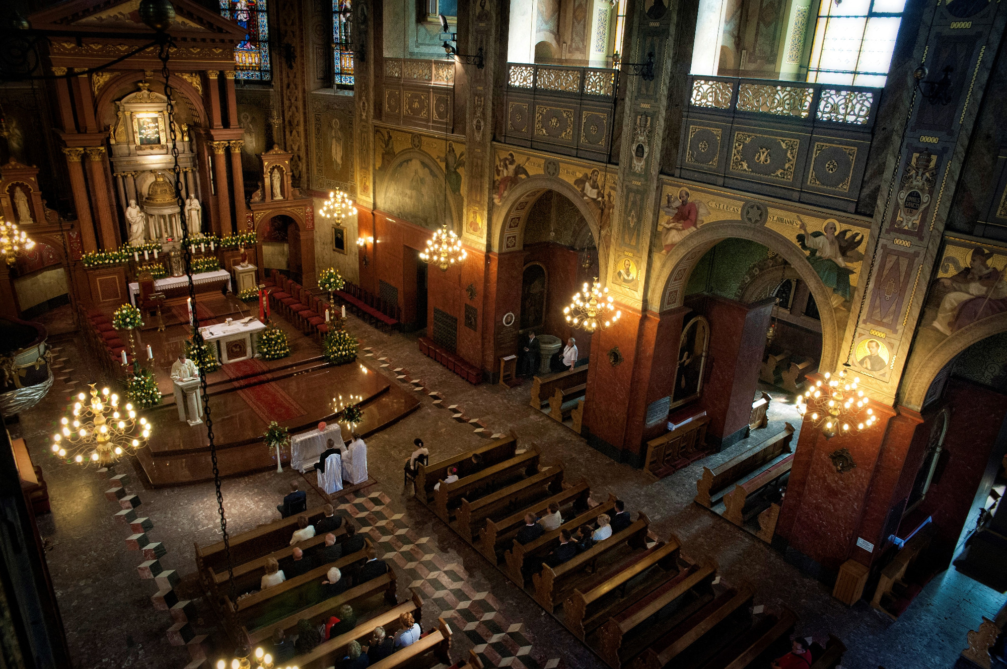 Wnętrze kościoła z widokiem z góry na ołtarz, księdza i parę młodą w trakcie ceremonii ślubnej, drewniane ławki wypełnione gośćmi, bogato zdobione ściany i sufit, oświetlone żyrandolami.
