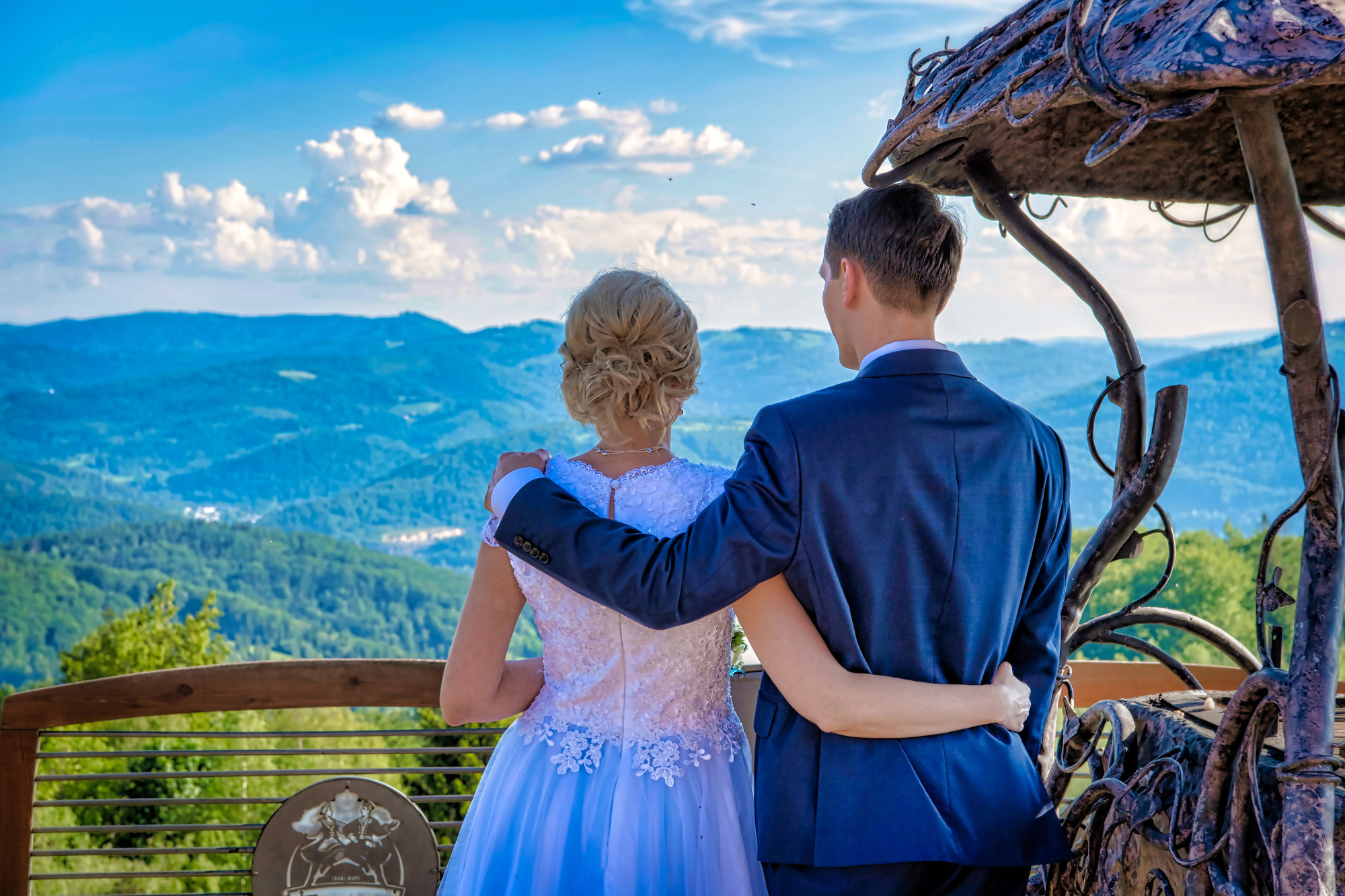 Para młoda, on w granatowym garniturze, ona w białej sukni z koronkowymi zdobieniami, stoi obejmując się na tle malowniczego krajobrazu górskiego z błękitnym niebem i chmurami, widok z altany.