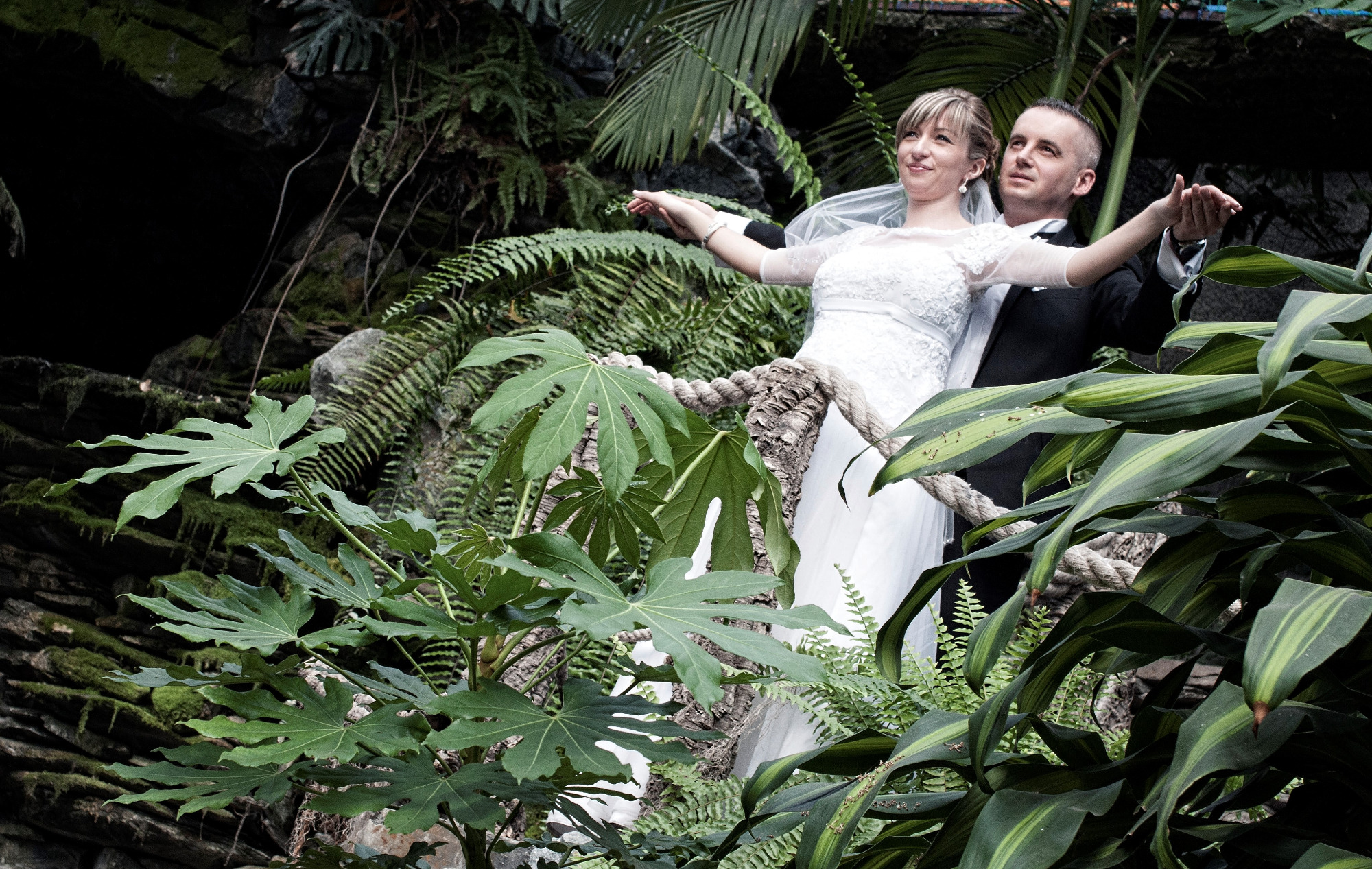 Para młoda w tropikalnej scenerii: panna młoda w białej sukni i welonie, pan młody w czarnym smokingu, pozują na tle bujnej roślinności i kamiennej ściany.