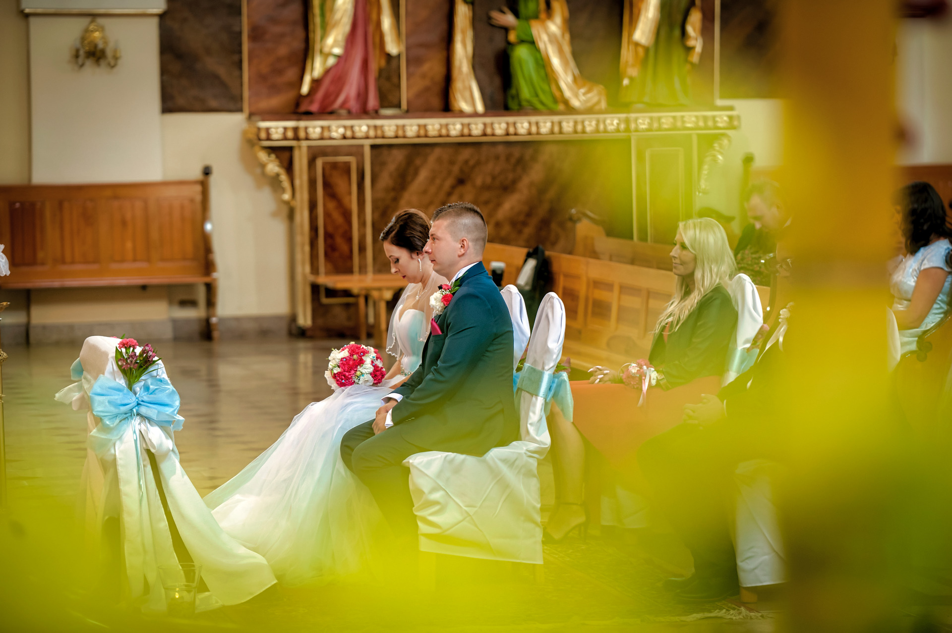 Para młoda siedzi w kościele podczas ceremonii ślubnej, widoczne dekoracje kwiatowe i ozdobne krzesła.
