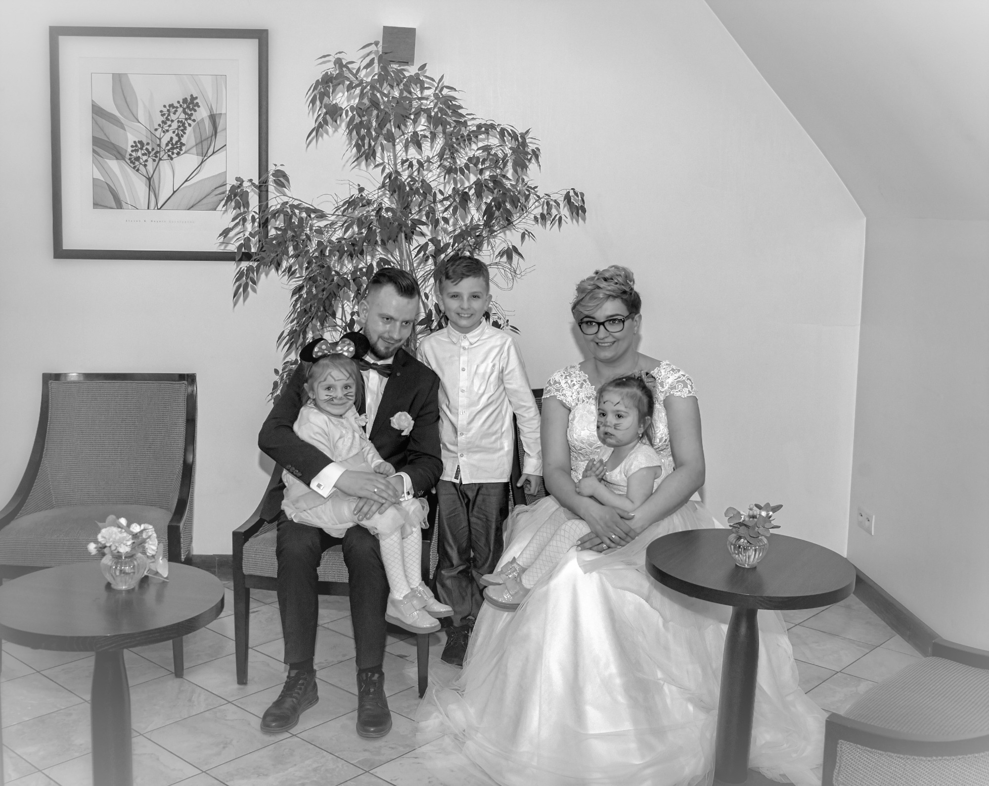 Czarno-białe zdjęcie rodziny: panna młoda w sukni ślubnej, pan młody w smokingu, starszy syn i dwie młodsze córki z pomalowanymi twarzami, siedzą na krzesłach w pomieszczeniu z rośliną doniczkową...