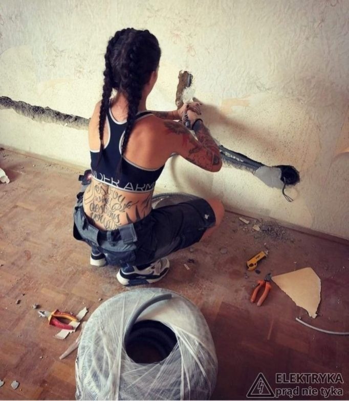 Kobieta w sportowym stroju, z tatuażami na plecach i ramionach, kuca przy ścianie i pracuje przy instalacji przewodów elektrycznych. Widoczne narzędzia, przewody w peszlu i fragmenty starej tapety...