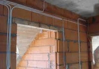 Surowe mury z cegły dziurawki z widocznymi, prowadzonymi natynkowo przewodami elektrycznymi w białych rurkach PCV, przed tynkowaniem.