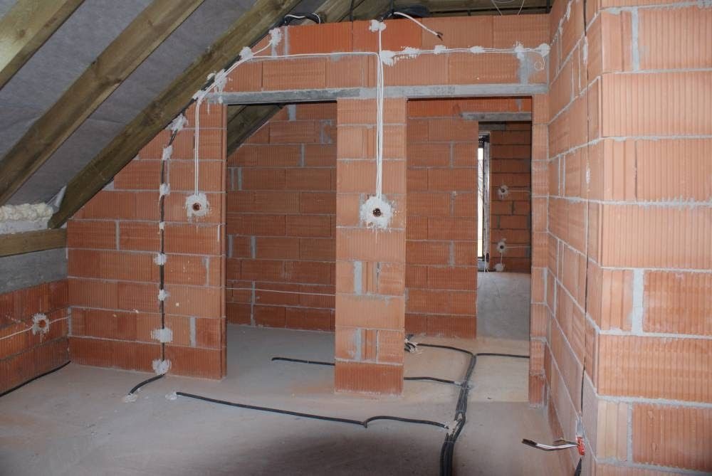 Wewnętrzne ściany z czerwonej cegły z widocznymi przewodami elektrycznymi i puszkami podtynkowymi, surowy stan wykończenia wnętrza poddasza.