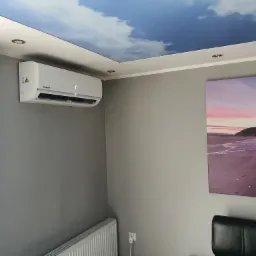 Biały klimatyzator Kaisai Eco zamontowany na szarej ścianie w pokoju z dekoracyjnym sufitem imitującym niebo, obok wiszą fioletowe zasłony i obraz z motywem plaży.