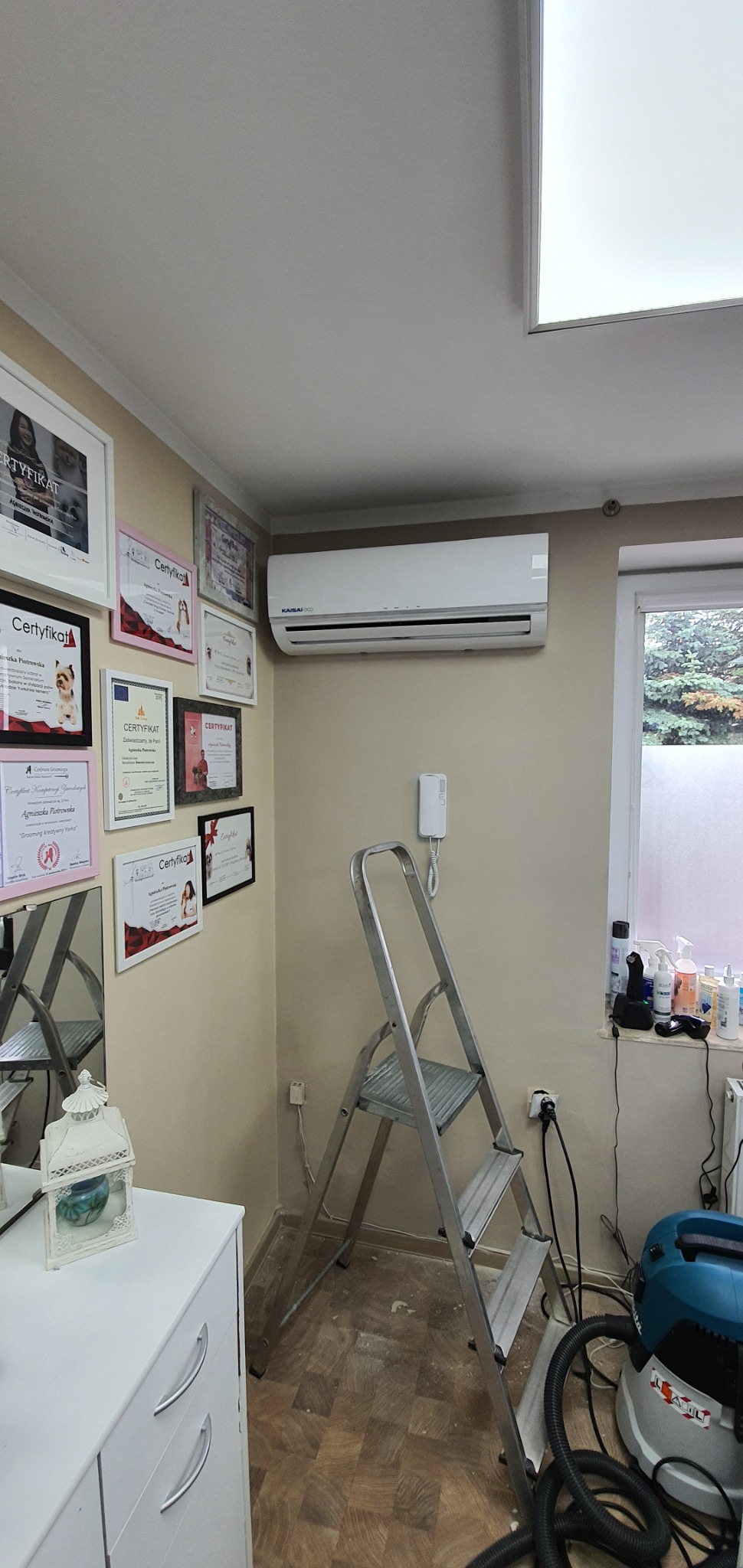 Wnętrze pomieszczenia z zamontowanym klimatyzatorem Kaisai ECO, drabina aluminiowa, odkurzacz przemysłowy oraz ściana z ramkami certyfikatów i dyplomów.