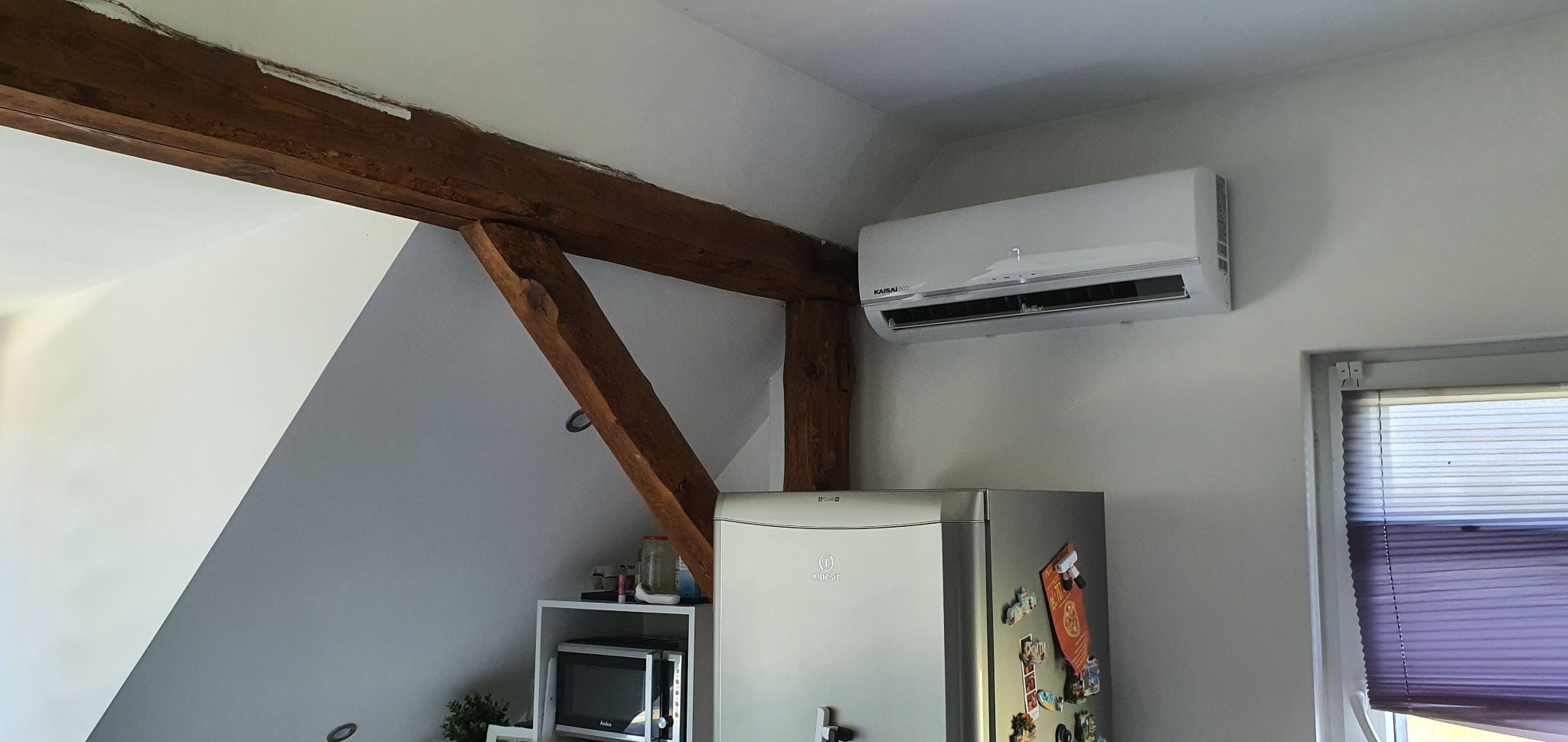 Biała jednostka klimatyzacji marki Kaisai zamontowana na ścianie nad lodówką Indesit, obok drewnianej belki stropowej w pomieszczeniu o skośnych ścianach.