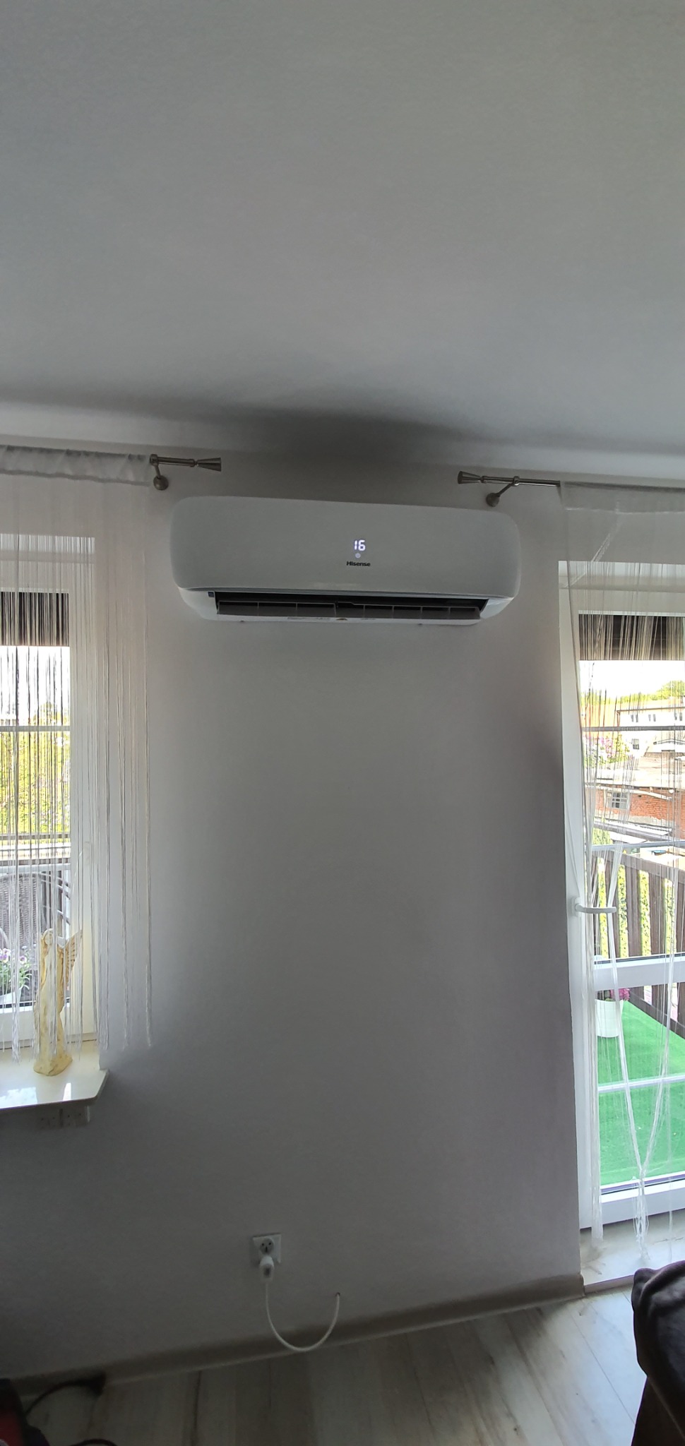 Biała jednostka klimatyzacji Hisense zamontowana na ścianie w pokoju w Bydgoszczy, widok z przodu, z wyświetlaną temperaturą 16 stopni Celsjusza, zasłony w oknach po bokach.