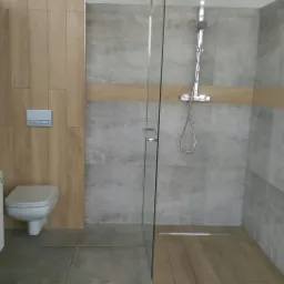 Wykończona łazienka z szarą płytką imitującą beton, drewnopodobną okładziną ścienną, kabiną prysznicową ze szklaną ścianką i odpływem liniowym, oraz białą szafką z umywalką i podwieszaną toaletą.