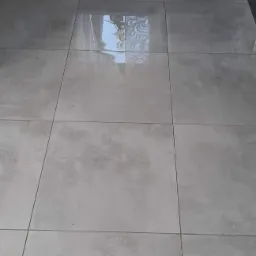 Ułożona podłoga z dużych, kwadratowych, szarych płytek z delikatnym wzorem i ciemnymi fugami, odbijająca światło, widoczny fragment czarnej szafki w tle.