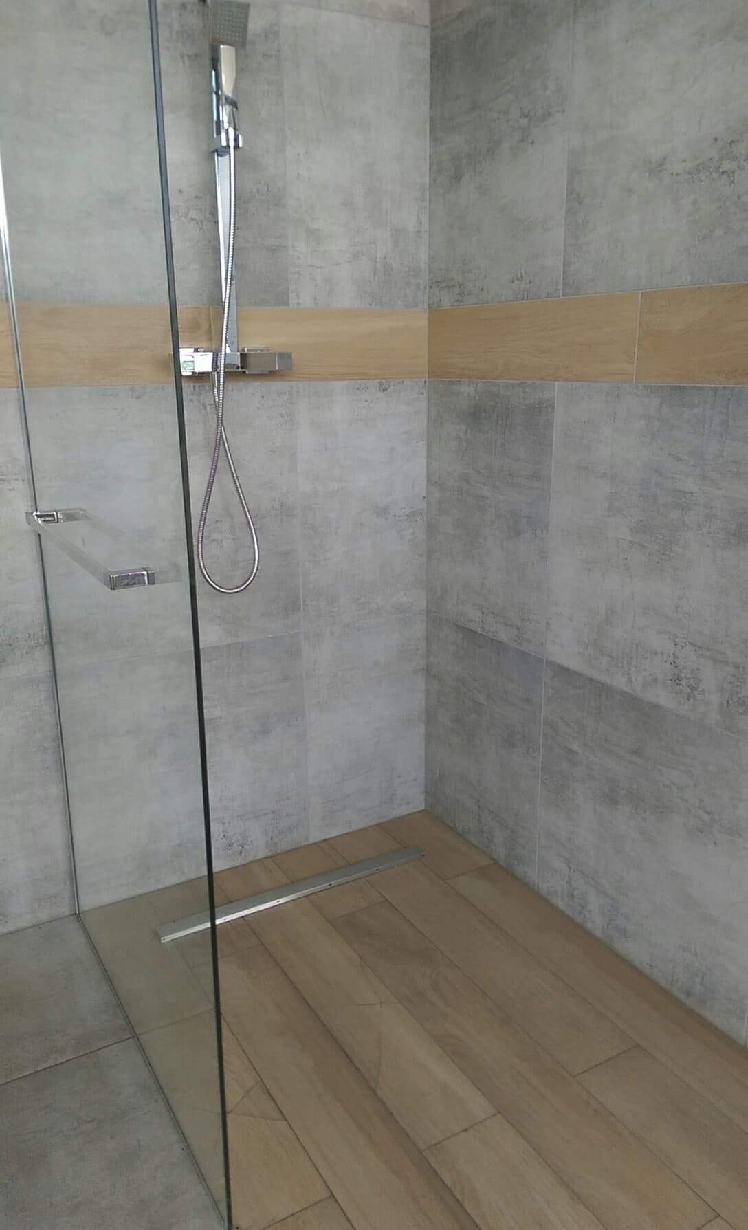 Kabina prysznicowa walk-in z odpływem liniowym, wykończona szarymi płytkami imitującymi beton i drewnopodobną mozaiką, chromowana bateria natryskowa.
