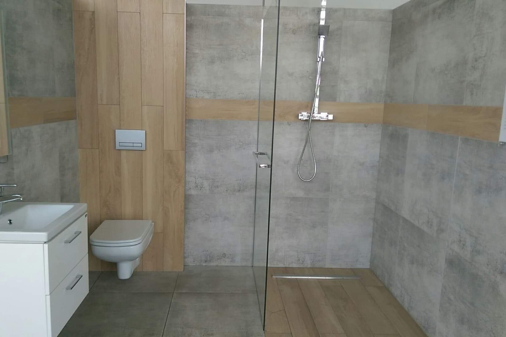 Wykończona łazienka z szarą płytką imitującą beton, drewnopodobną okładziną ścienną, kabiną prysznicową ze szklaną ścianką i odpływem liniowym, oraz białą szafką z umywalką i podwieszaną toaletą.