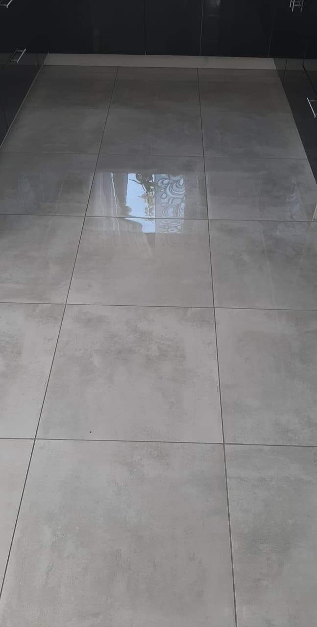 Ułożona podłoga z dużych, kwadratowych, szarych płytek z delikatnym wzorem i ciemnymi fugami, odbijająca światło, widoczny fragment czarnej szafki w tle.