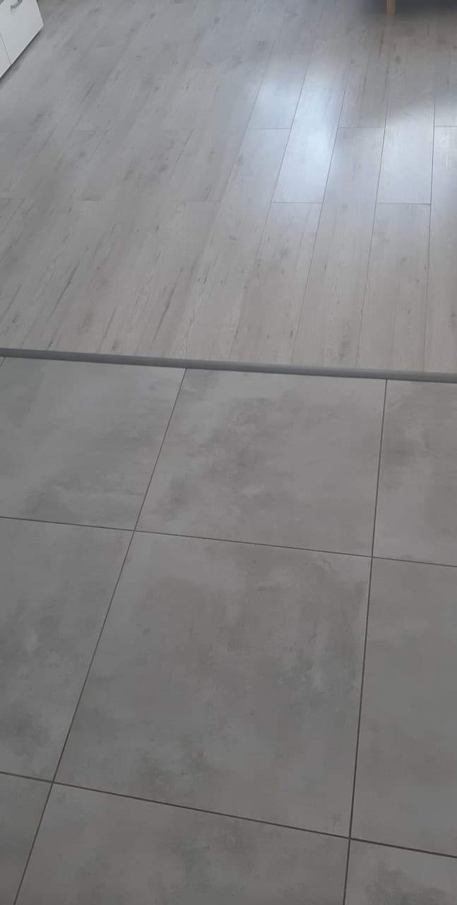 Połączenie jasnych paneli podłogowych i szarych, matowych płytek podłogowych z szarą listwą dylatacyjną.