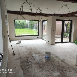 k-nest constructions Karol Wit - Wnętrze w trakcie prac wykończeniowych: ściany po nałożeniu gładzi, widoczne instalacje pod sufitem, podłoga zabezpieczona przed zabrudzeniem, okno z widokiem na ogród, wiadro z masą szpachlową.