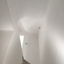 Wnętrze w trakcie remontu: schody z surowego betonu kontrastują z gładkimi, białymi ścianami i sufitem. Widoczne ślady po pracach wykończeniowych oraz wystający przewód elektryczny. Minimalistyczne...