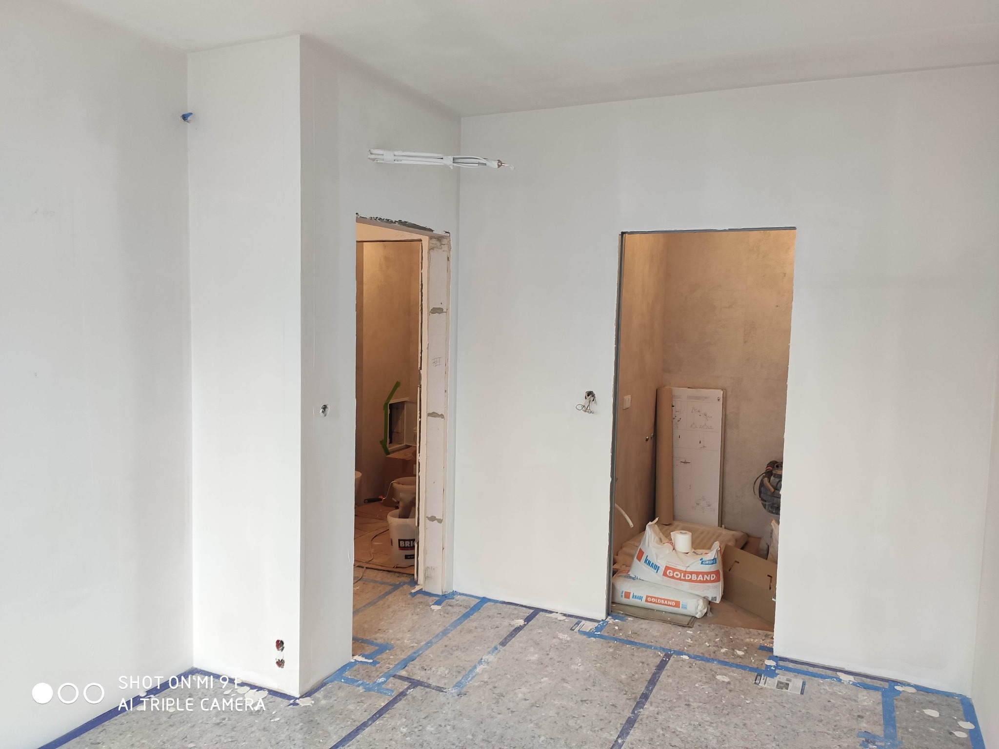 Wnętrze po remoncie, ściany świeżo pomalowane na biało, widoczne worki z gipsem budowlanym Knauf Goldband, podłoga zabezpieczona taśmą malarską, instalacja elektryczna pod sufitem czeka na zabudowę.