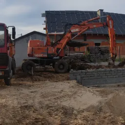 Pomarańczowa koparka Atlas 1304K pracuje przy wykopie pod fundamenty domu z cegły, obok stoi czerwona ciężarówka Mercedes, w tle konstrukcja dachu.