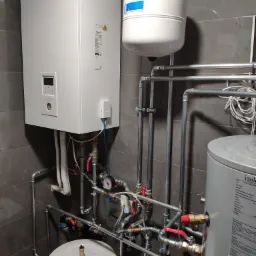 Biała jednostka wewnętrzna Panasonic z podłączonymi rurami, zbiornik wyrównawczy i zasobnik ciepłej wody w kotłowni wykończonej płytkami imitującymi kamień.