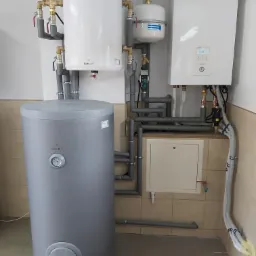 Kompletna instalacja grzewcza z szarym zasobnikiem Galmet, białym zbiornikiem buforowym i kotłem grzewczym Panasonic, połączona rurami w pomieszczeniu z beżowymi płytkami.