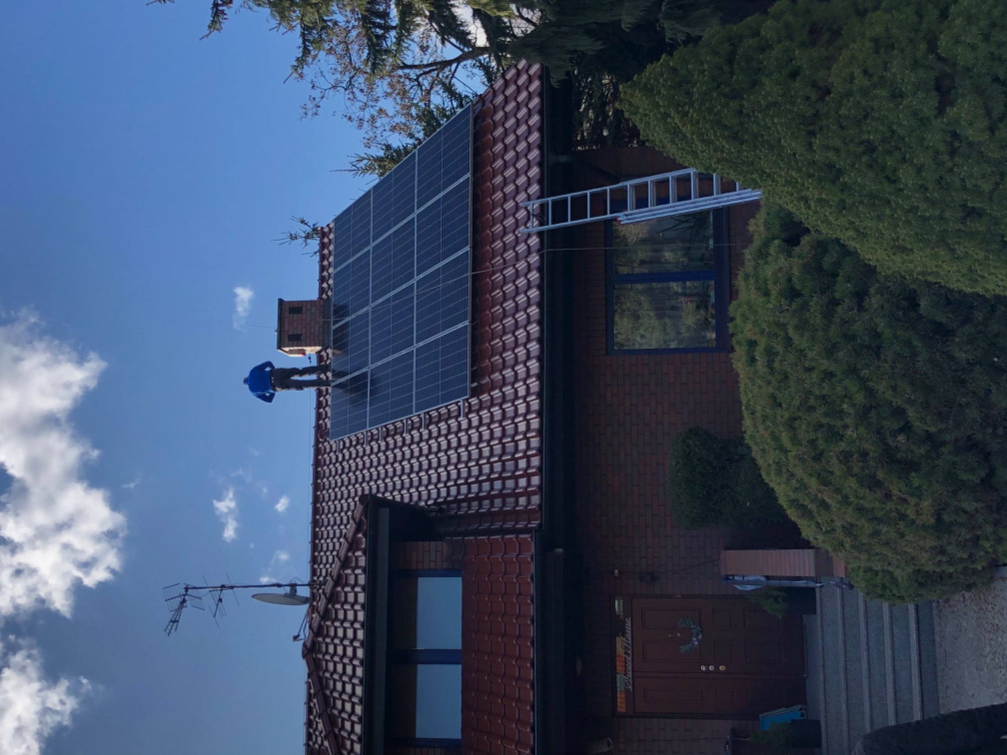 Instalator w niebieskiej kurtce na dachu z czerwonej dachówki, montujący panele słoneczne; drabina oparta o ścianę budynku z cegły; w tle błękitne niebo z chmurami.