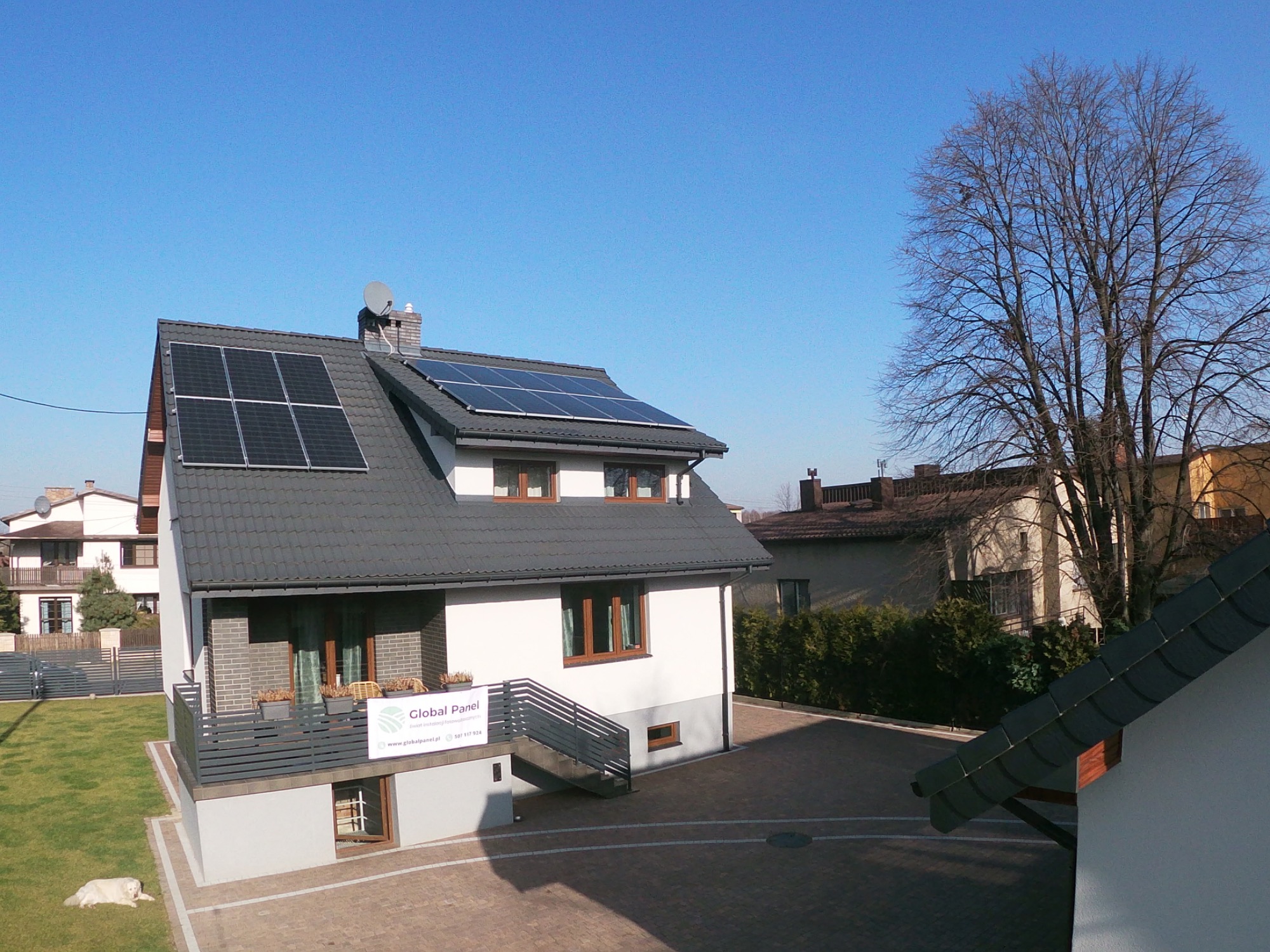 Dom jednorodzinny z szarym dachem, na którym zamontowano panele fotowoltaiczne, widoczny z boku, z psem leżącym na trawniku i banerem reklamowym firmy na balkonie.