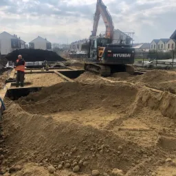 Żółty piasek usypany w wykopie pod fundamenty domu jednorodzinnego, w tle pracuje koparka Hyundai, a obok stoi mężczyzna w pomarańczowej kamizelce.