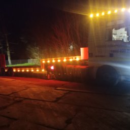 Bud-Car SP. z o.o., Sp. k. - Ciężarówka z naczepą niskopodwoziową, oświetlona lampami, stoi na placu w nocy. Widoczne drzewa w tle.