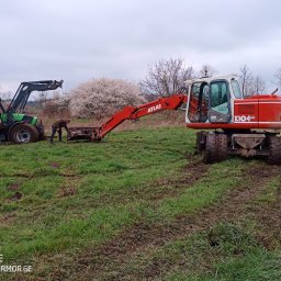 Bud-Car SP. z o.o., Sp. k. - Czerwony koparko-ładowarka Atlas 1304 i zielony traktor w trakcie robót ziemnych na podmokłym terenie z kwitnącymi drzewami w tle, operator przyczepia linę do ładunku.