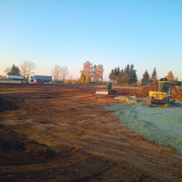 Bud-Car SP. z o.o., Sp. k. - Dwa żółte walce drogowe pracują na rozległym, wyrównanym terenie budowy o poranku, z widocznym świeżo rozłożonym kruszywem i śladami opon w ziemi, w tle drzewa i pojazdy.