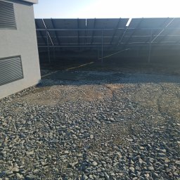 Bud-Car SP. z o.o., Sp. k. - Teren utwardzony szarym gruzem przy budynku z żaluzjami wentylacyjnymi, w tle konstrukcja paneli słonecznych.